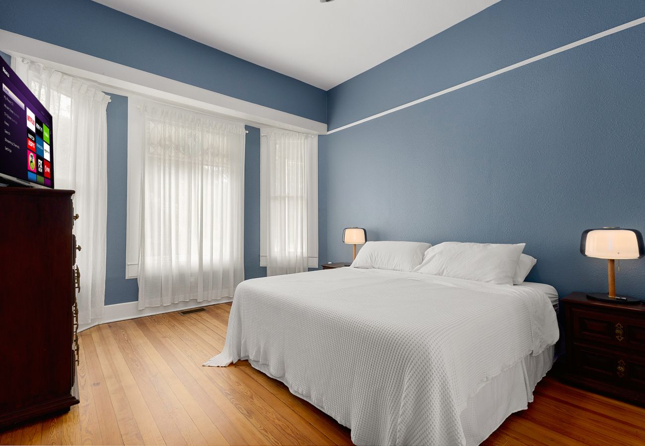 Primary Bedroom w/ Tall Windows & Smart TV | Seguin TX  Primary bedroom with wood floors, white bedding, bedside lamps, and smart TV in Seguin rental